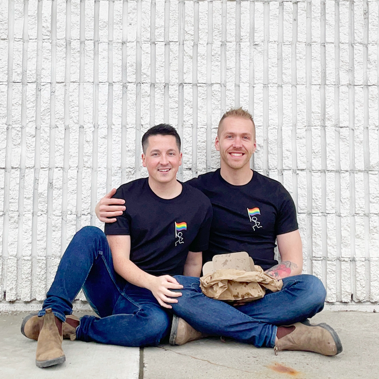 Two white gay men sitting on a sidewalk infront of a white wall wearing jeans, boots and matching t-shirts from Outtire. They're holding a box of fresh fries from a chip truck.